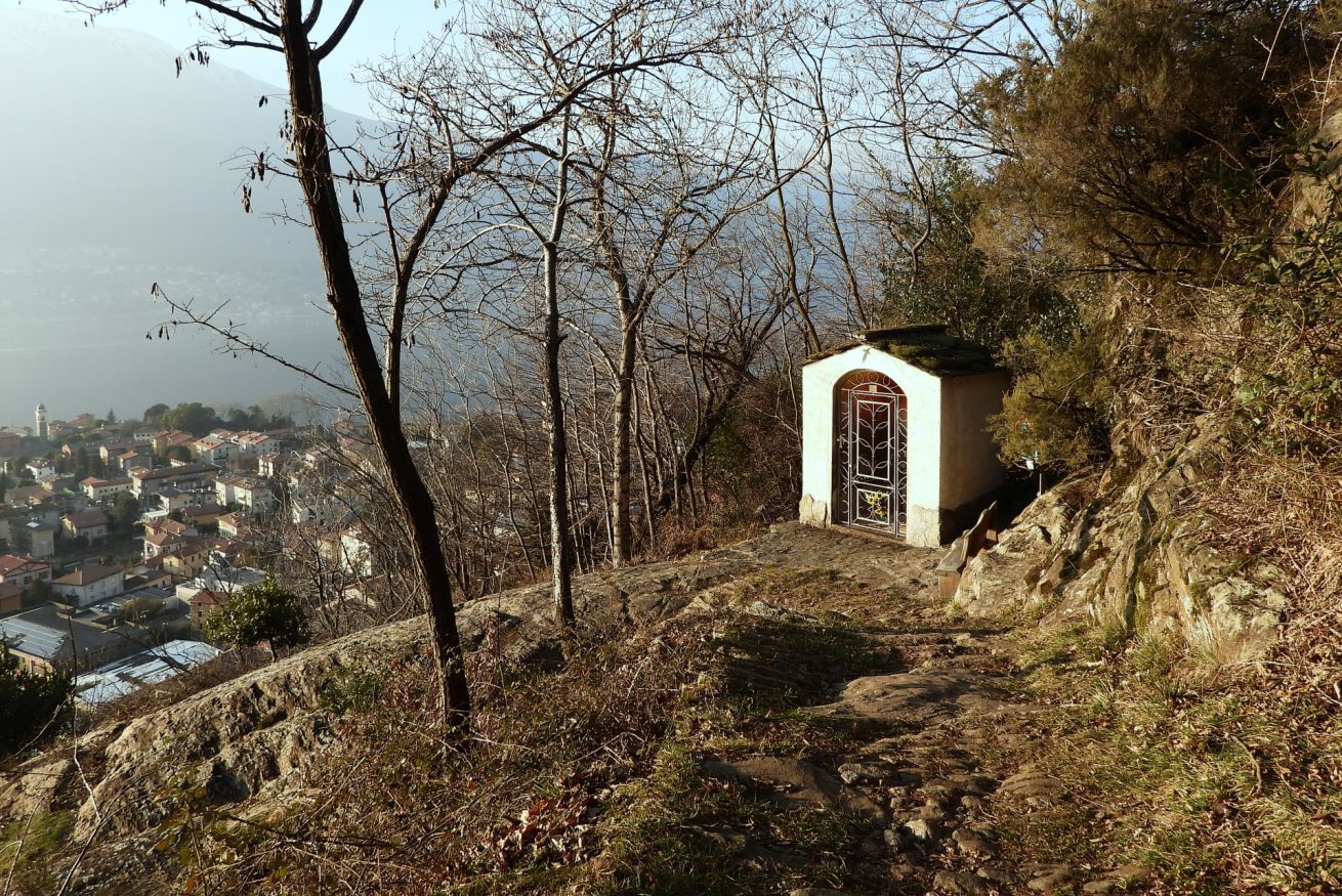 Una piccola cappella con facciata bianca e un portone, circondata da alberi e terreno roccioso, con vista su un paese.