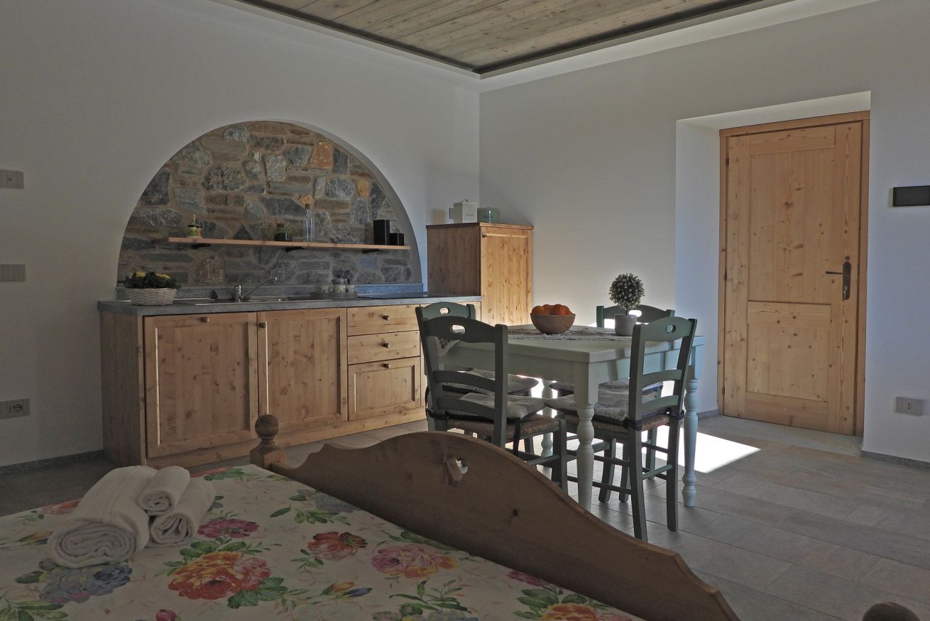 Une chambre à coucher avec un lit, une tête de lit en bois, une table à manger avec des chaises, un coin cuisine avec une arche en pierre et une porte en bois.