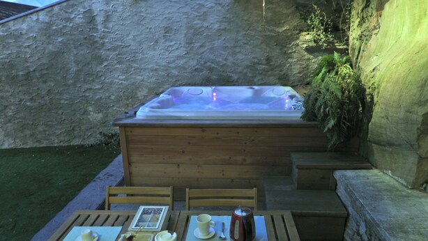 A hot tub with a wooden deck and steps leading up to it, surrounded by a stone wall and greenery.