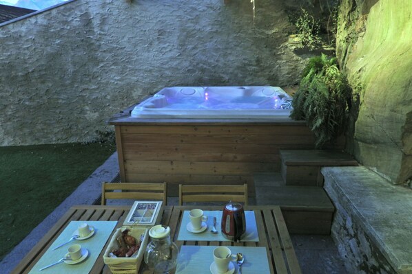 Un jacuzzi con cubierta de madera y escalones que llevan a él, rodeado de un muro de piedra y follaje.