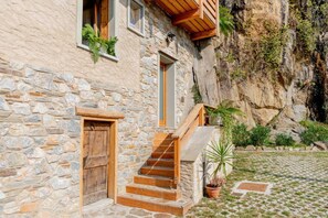 A stone building with a wooden staircase leading to a door, surrounded by greenery and a rocky cliff. - Green Chalet Scalotta - private Jacuzzi and SPA - few steps from lake and best trekking (Dervio)