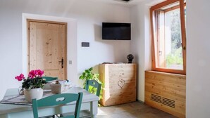 A dining area with a table, chairs, a TV mounted on the wall, a wooden cabinet with a heart design, and a window with a view of greenery. - Green Chalet Scalotta - private Jacuzzi and SPA - few steps from lake and best trekking (Dervio)