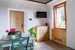 Ein Essbereich mit Tisch, Stühlen, einem an der Wand montierten Fernseher, einem Holzschrank mit Herzmotiv und einem Fenster mit Blick auf Grünflächen.