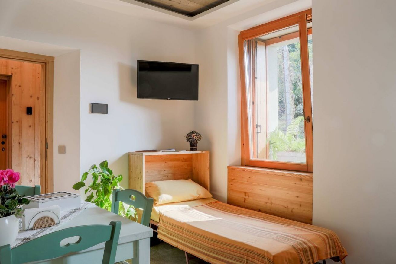 Une petite pièce agréable et chaleureuse, avec un lit simple, une table et des chaises, un téléviseur fixé au mur et une fenêtre laissant entrer la lumière naturelle.