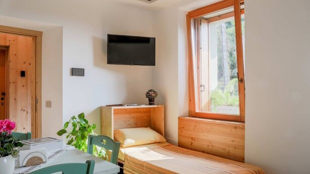 A small, cozy room with a single bed, a table with chairs, a TV mounted on the wall, and a window providing natural light. - Green Chalet Scalotta - private Jacuzzi and SPA - few steps from lake and best trekking (Dervio)