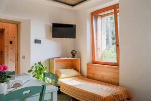 Une petite pièce agréable et chaleureuse, avec un lit simple, une table et des chaises, un téléviseur fixé au mur et une fenêtre laissant entrer la lumière naturelle.