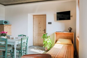 Una habitación con puerta de madera, una cama con manta a rayas, un televisor montado en la pared, una mesa de comedor con sillas y una planta en maceta.