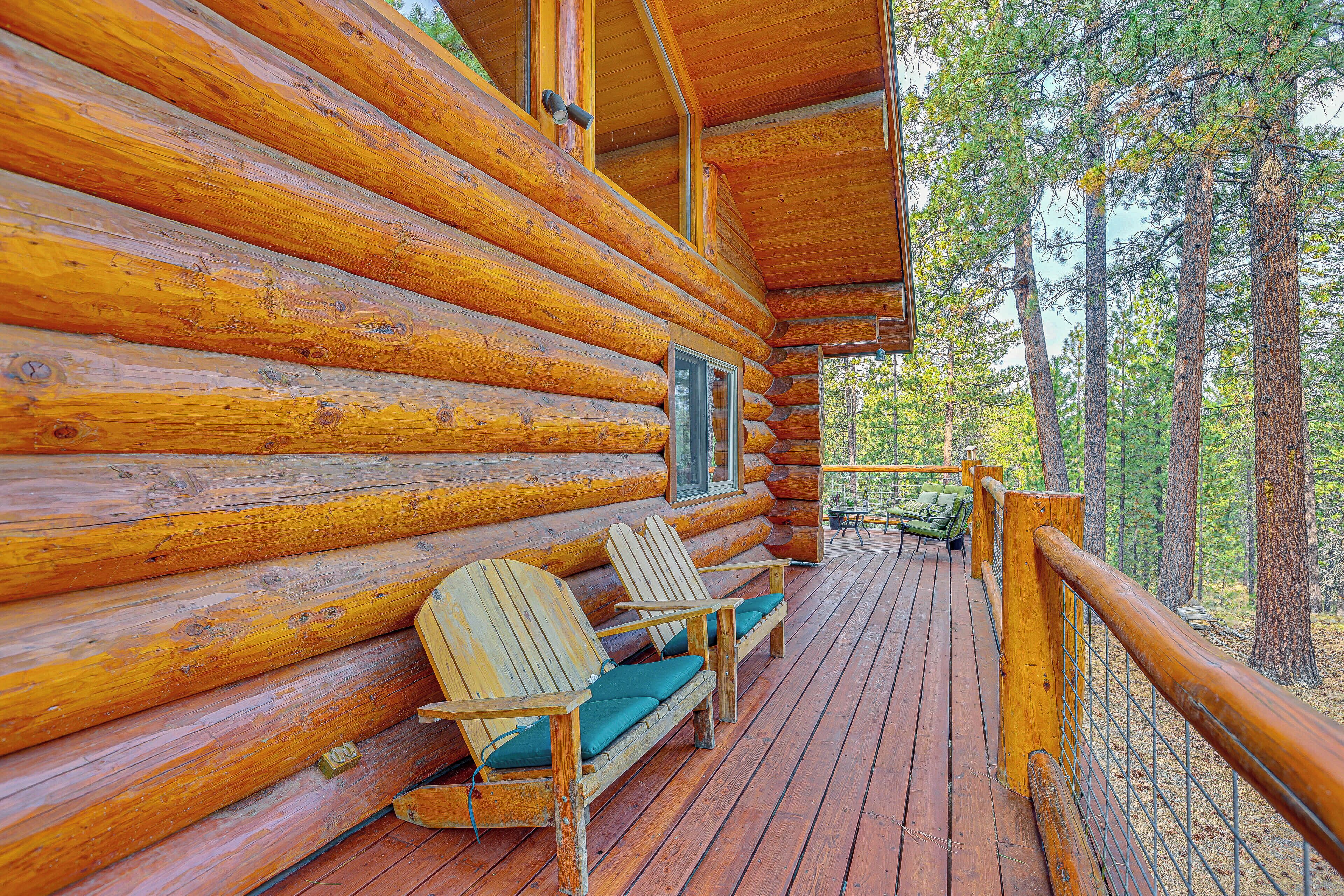 A log cabin with a wooden deck, two Adirondack chairs, and a view of the forest.
