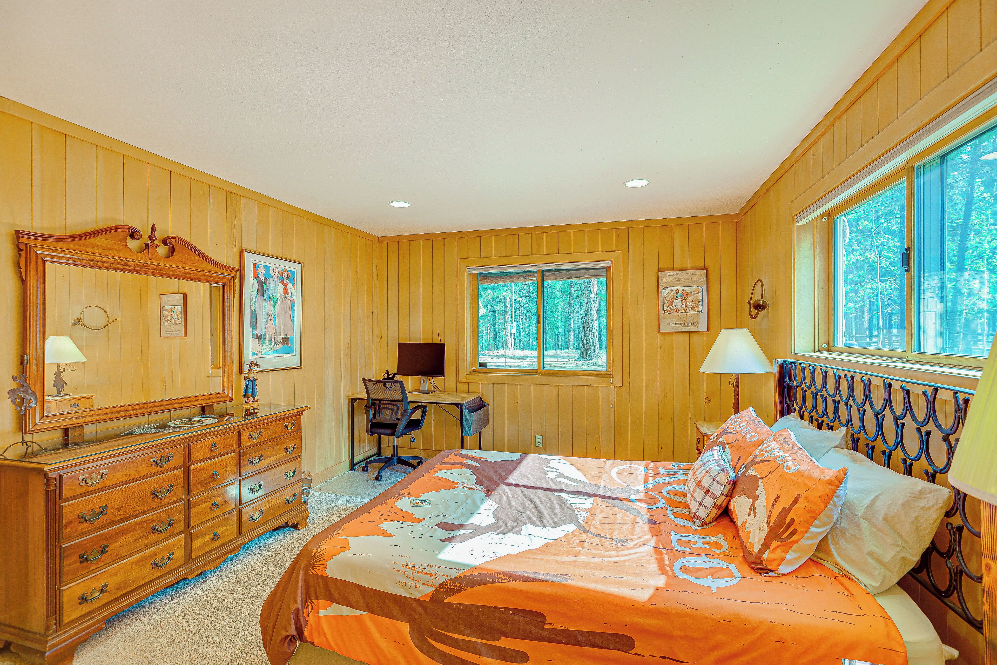 A bedroom with a wooden paneled wall, a large bed with an orange bedspread, a dresser, a desk with a computer, and a window with a view of trees.