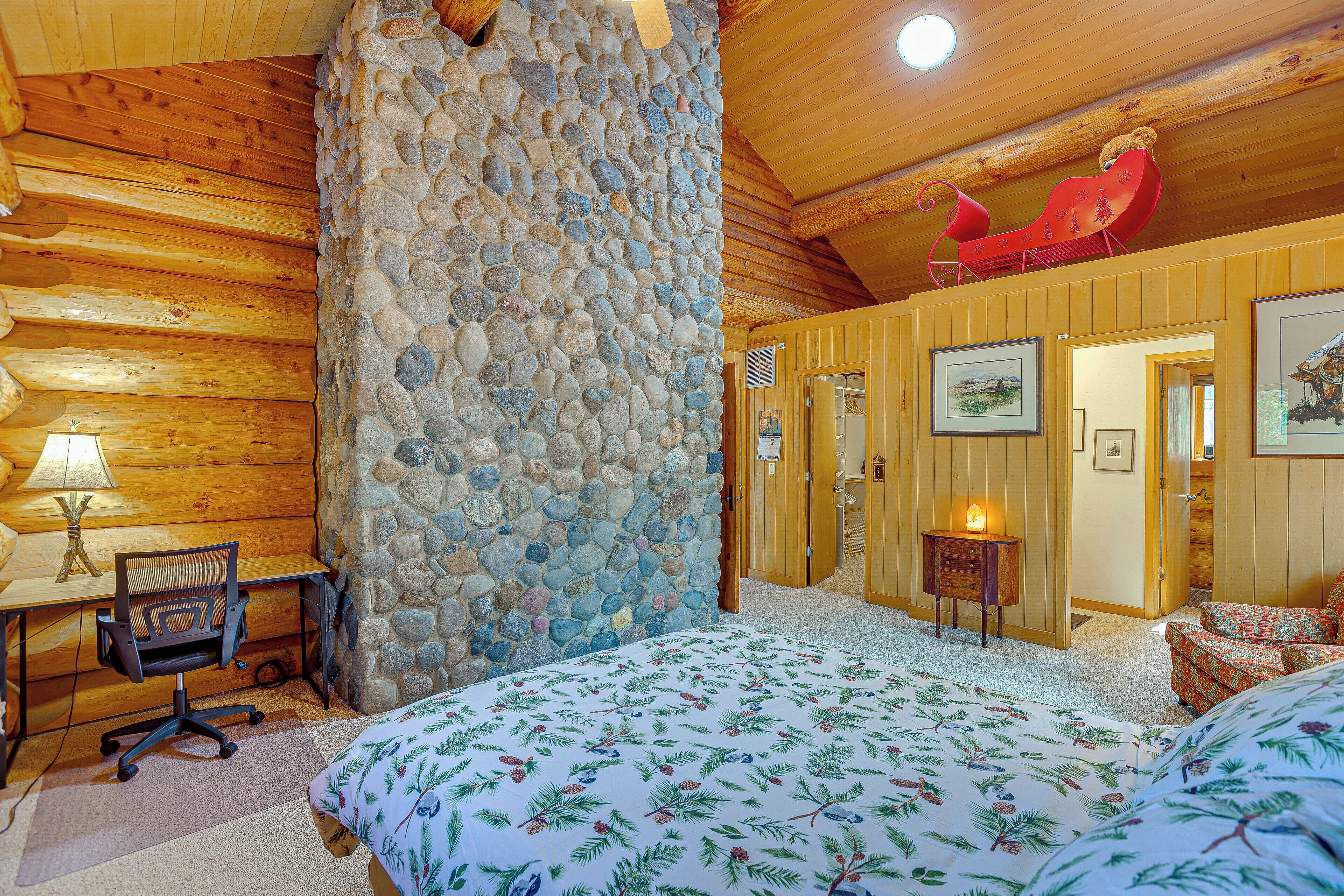 A cozy log cabin room with a stone wall, wooden ceiling, a bed with a floral bedspread, a desk with a chair, and a red sleigh hanging from the ceiling.