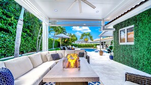 An outdoor living area with a fire feature, seating, and a view of a pool and greenery. - Heated SaltWater Pool+HotTub! Golf Cart+1 Mile To Beach! (Fort Lauderdale)