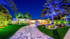 A well-lit pathway leading to a house with a stone patio, surrounded by lush greenery and palm trees. - Heated SaltWater Pool+HotTub! Golf Cart+1 Mile To Beach! (Fort Lauderdale)