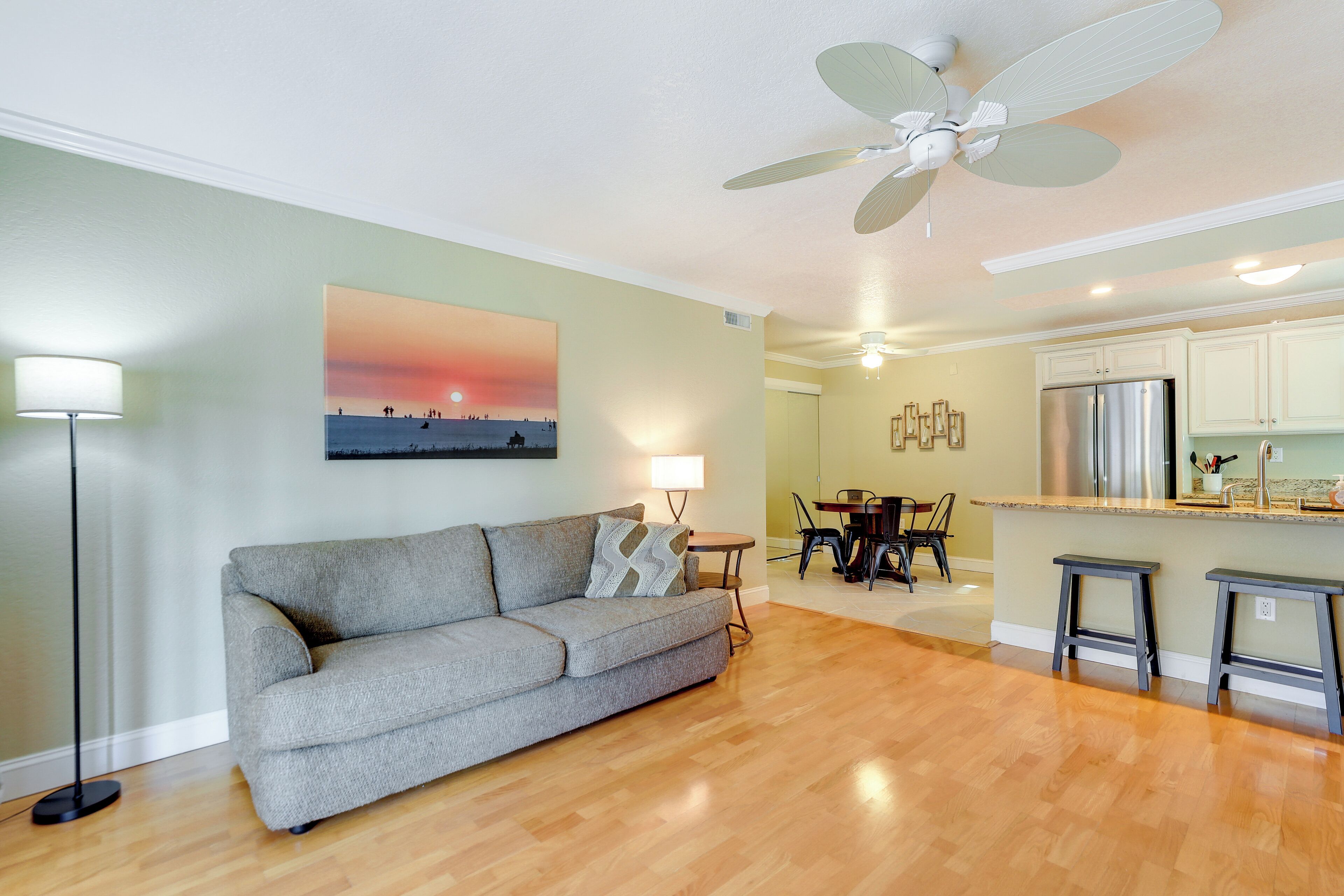 A living room with a gray sofa, a ceiling fan, a painting of a sunset, a dining area with a table and chairs, and a kitchen with a refrigerator.
