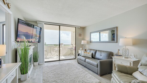 A modern living room with a grey sofa, a white coffee table, and a TV mounted on the wall. There is a sliding glass door leading to an outdoor balcony with a table and chairs. - Gulf-view Balcony & Pool Access: Hudson Retreat (Hudson)