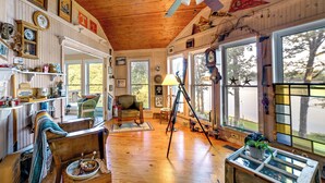 A cozy cabin interior with wooden floors, a ceiling fan, a rocking chair, a tripod, and large windows offering a view of the outdoors. - Pet-friendly New Concord Vacation Rental on Lake! (New Concord)