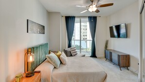 A modern bedroom with a bed, a ceiling fan, a TV, and a window with curtains. - Luxe Resort Condo - 2 Mi to Daytona Beach! (Holly Hill)