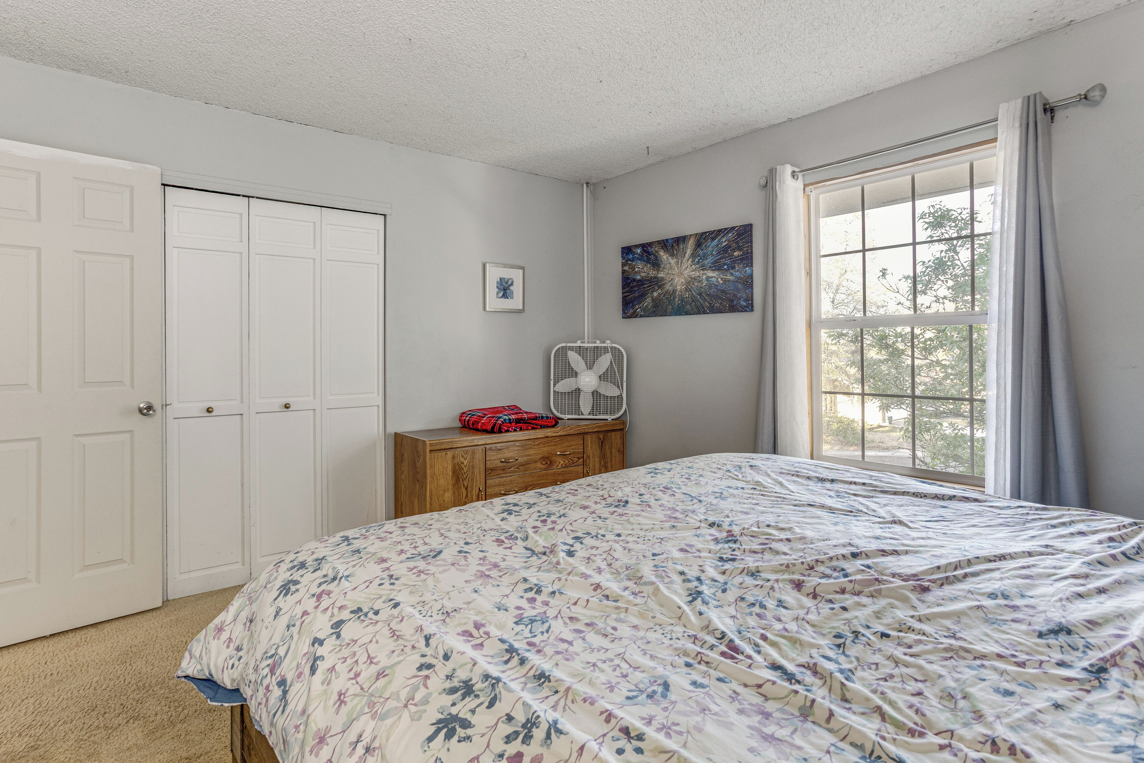 A bedroom with a bed, a dresser, a fan, a window with curtains, and a door.
