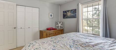 A bedroom with a bed, a dresser, a fan, a window with curtains, and a door.
