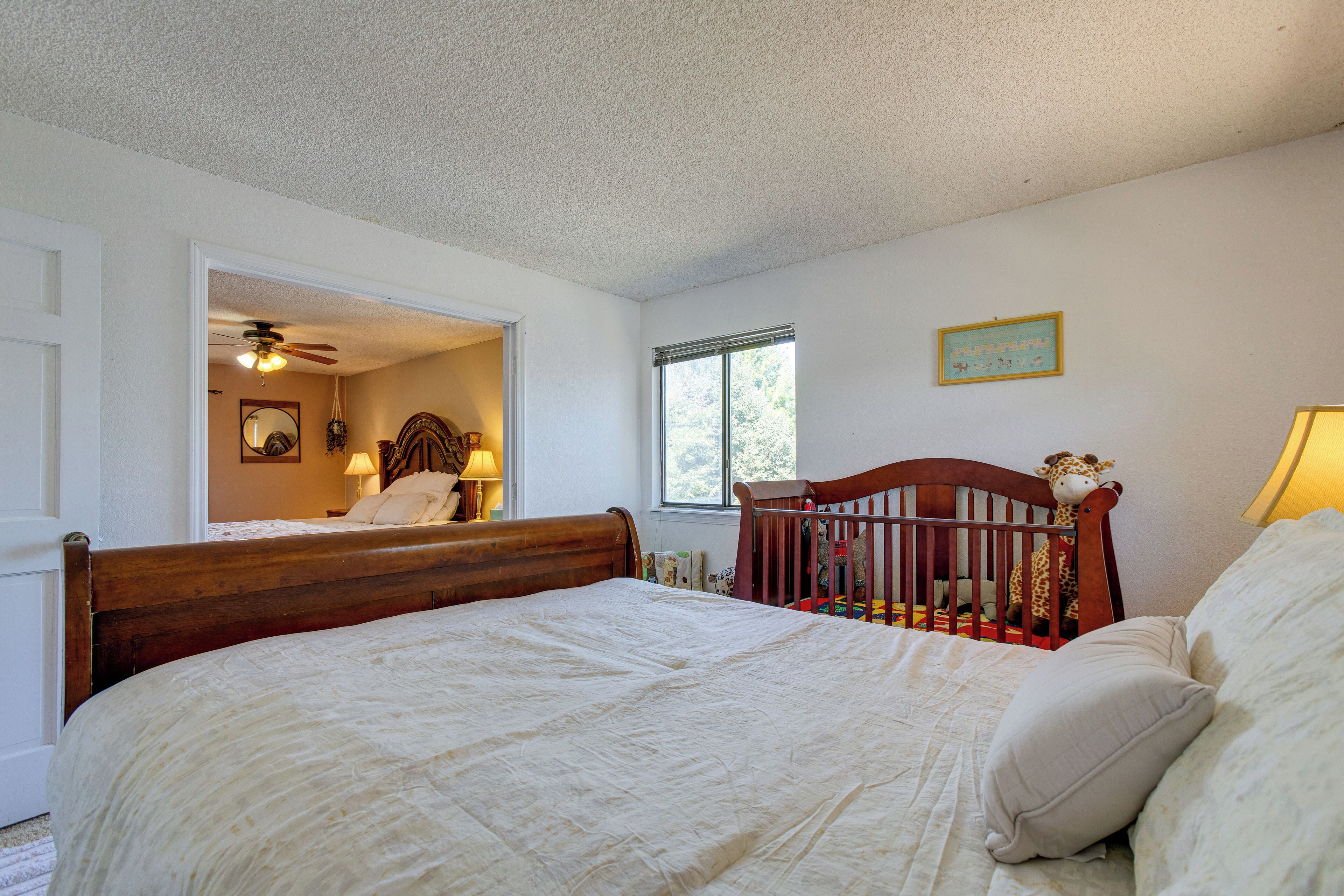 A bedroom with a wooden bed, a crib with a giraffe toy, a window with a view, and a ceiling fan.