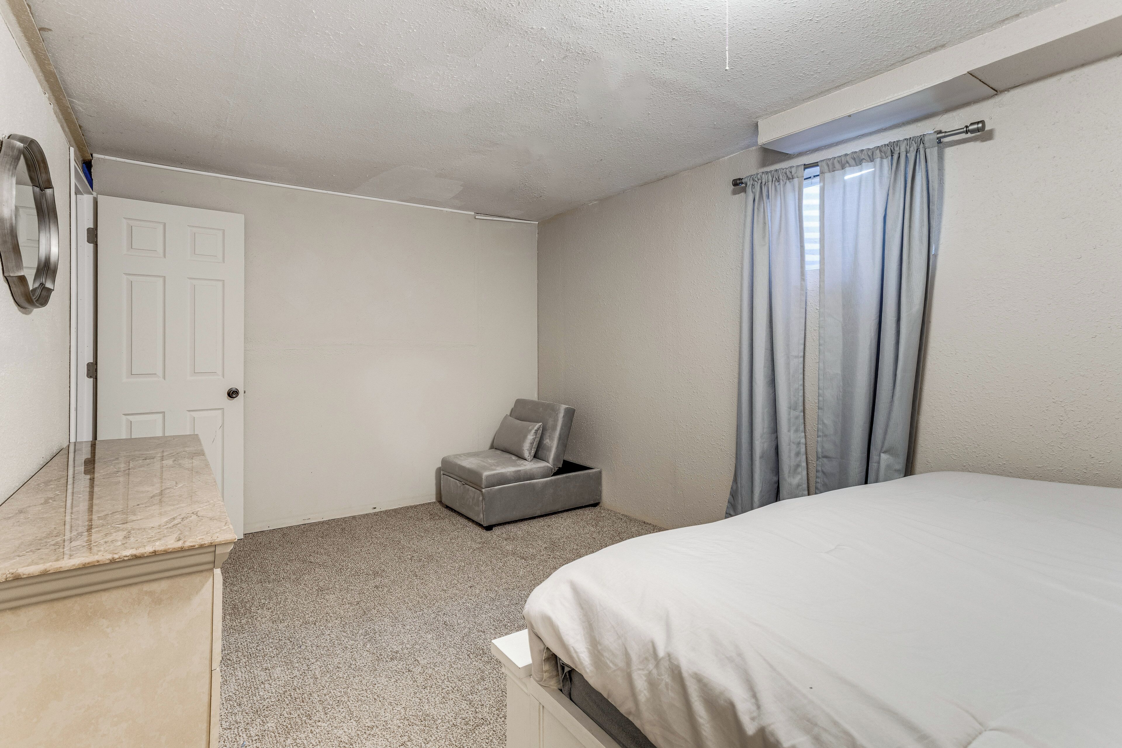 A bedroom with a bed, a small seating area, a door, and a window with curtains.