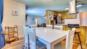 Un comedor amplio con una mesa y sillas blancas, suelo de madera y una cocina con armarios de madera y una campana extractora de acero inoxidable.