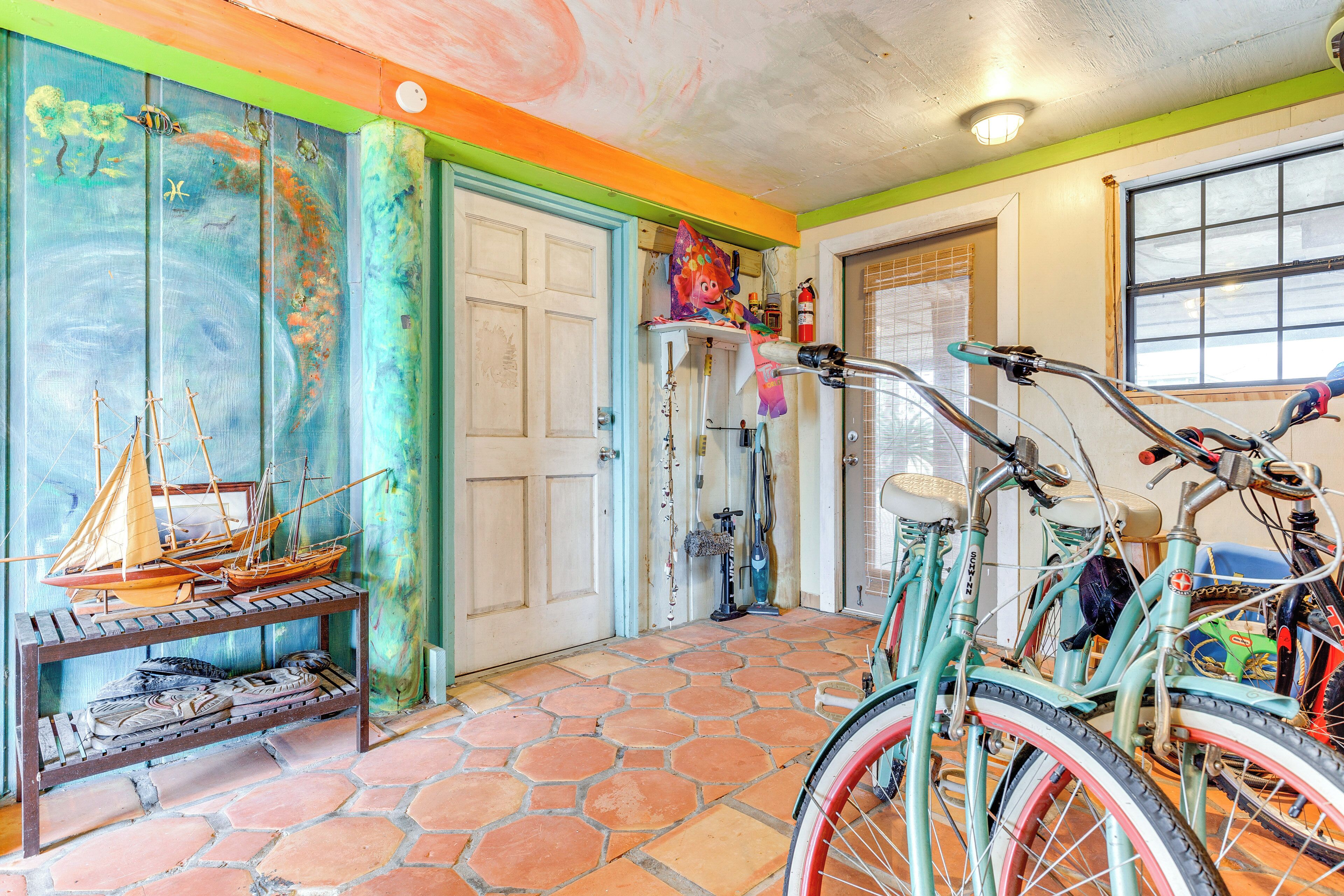 Une pièce avec un sol carrelé, une étagère en bois où sont rangées des chaussures, un vélo et une porte ornée d’une fresque colorée.