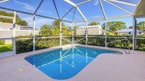A covered swimming pool with a clear blue water surface, surrounded by a metal frame structure, and a view of residential buildings in the background. - Charming St Lucie River Retreat w/ Pool & Dock! (Port Saint Lucie)