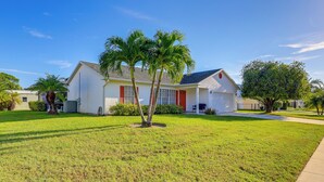A single-story house with a red door and white walls, surrounded by a well-maintained lawn and palm trees. - Charming St Lucie River Retreat w/ Pool & Dock! (Port Saint Lucie)