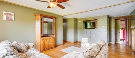 A spacious living room with a large sectional sofa, wooden cabinet, ceiling fan, and a flat-screen TV mounted on the wall.