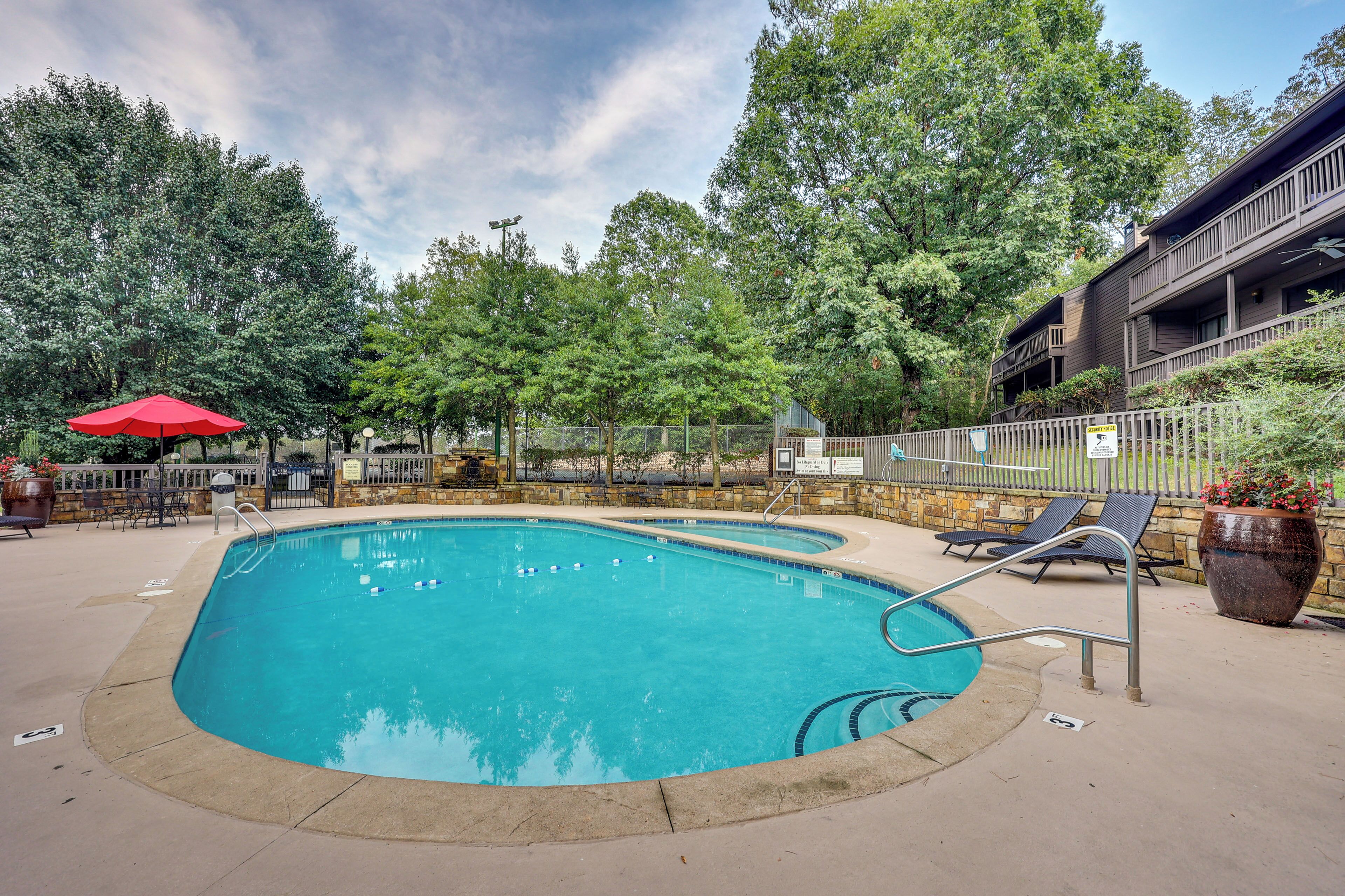 A rectangular swimming pool with a metal ladder, surrounded by a concrete deck, with lounge chairs and a red umbrella.