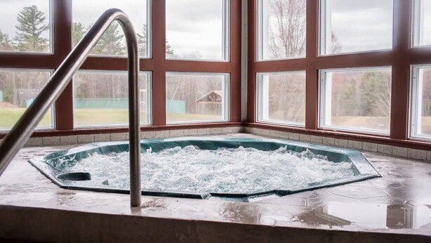 Un jacuzzi con una escalera metálica frente a un ventanal con vistas a árboles y un edificio.