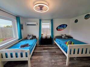 Quarto com duas camas de solteiro, ventilador de teto, aparelho de ar condicionado, janela com cortinas e um adorno na parede.