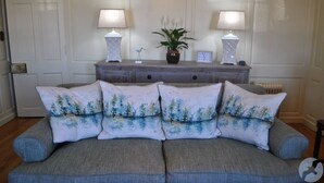 Sala de estar com sofá cinza, almofadas florais, mesa lateral de madeira com duas luminárias, planta em vaso e um relógio.