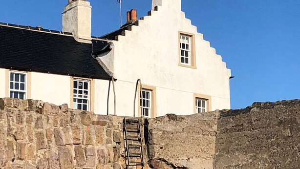 Um edifício branco com várias chaminés e janelas, uma parede de pedra com uma escada, e um céu azul claro.
