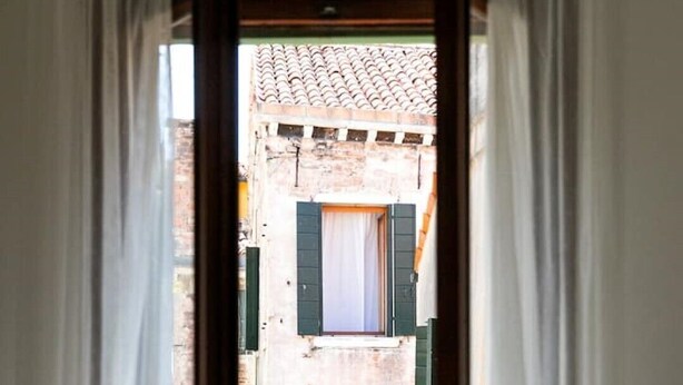 A view through a doorway with white curtains, leading to a narrow street with buildings featuring exposed brickwork and green shutters. - In the Art Quartier (Venezia)