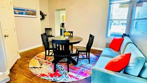 A dining area with a round table, chairs, and a colorful rug. There is a blue sofa with red cushions and a window with blinds.