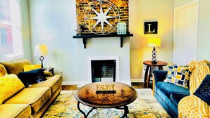 A living room with a brick fireplace, a round coffee table with 'ENJOY' on it, a blue sofa, and a yellow armchair.
