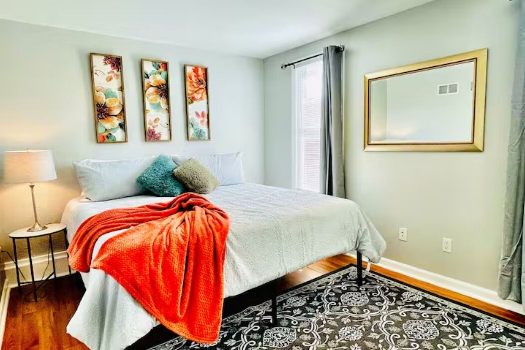 A bedroom with a bed, a nightstand, a lamp, a window with curtains, and three wall-mounted paintings.