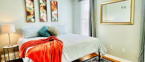 A bedroom with a bed, a nightstand, a lamp, a window with curtains, and three wall-mounted paintings.