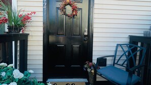 A black front door with a wreath, a 'hello' mat, and a chair outside.