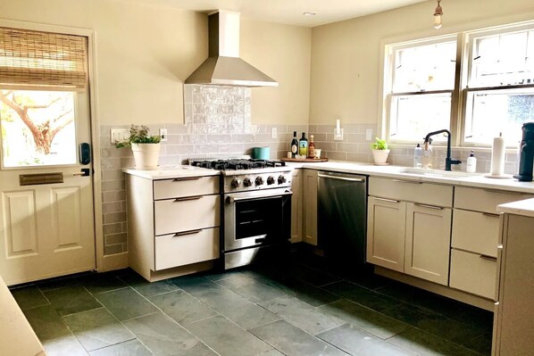 A compact kitchen with a gas stove, oven, and dishwasher. It features light-colored cabinetry, a tiled backsplash, and a window with blinds. - Charming Hydrangea Cottage in Sewickley Village 4bd/1.5 b - 20 min to Pittsburgh (Sewickley)