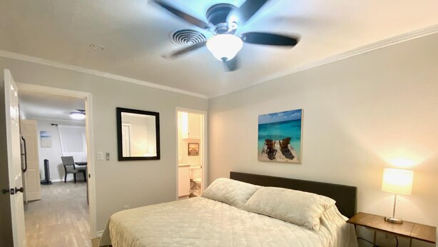 Een slaapkamer met een bed, een plafondventilator, een nachtkastje met een lamp en een schilderij van een strandgezicht.