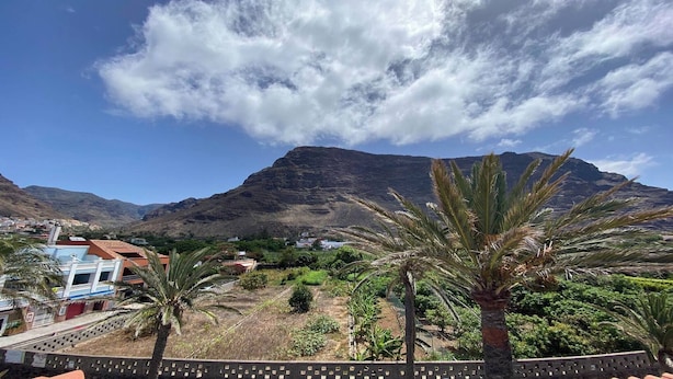 Paisaje montañoso con cielo despejado, algunos edificios y palmeras.