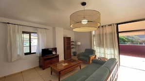 Una sala de estar con un sofá, un televisor sobre un mueble de madera, una lámpara de techo de mimbre, un ventilador y vistas a un balcón con barandilla.