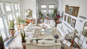 Un salón espacioso con mobiliario blanco, ventanas amplias y varias plantas en macetas.