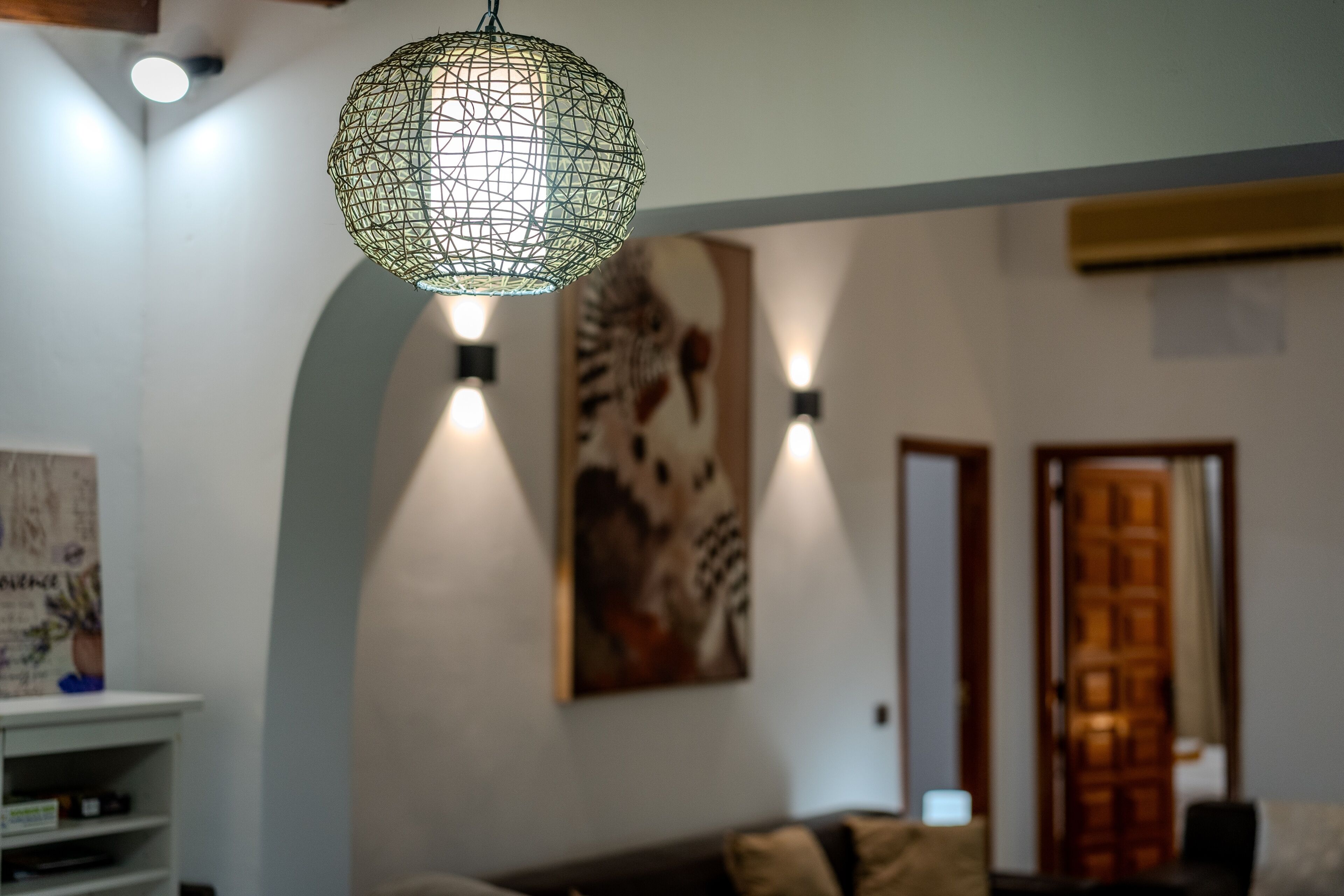 A room with a geometric pendant light, a sofa with cushions, a wall-mounted artwork, and a door with a wooden panel.