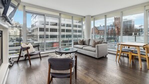 A spacious living room with a sofa, chairs, and a table, large windows offering a city view. - Waterfalls Hotel - Kings Corner - Resort Living (Victoria)