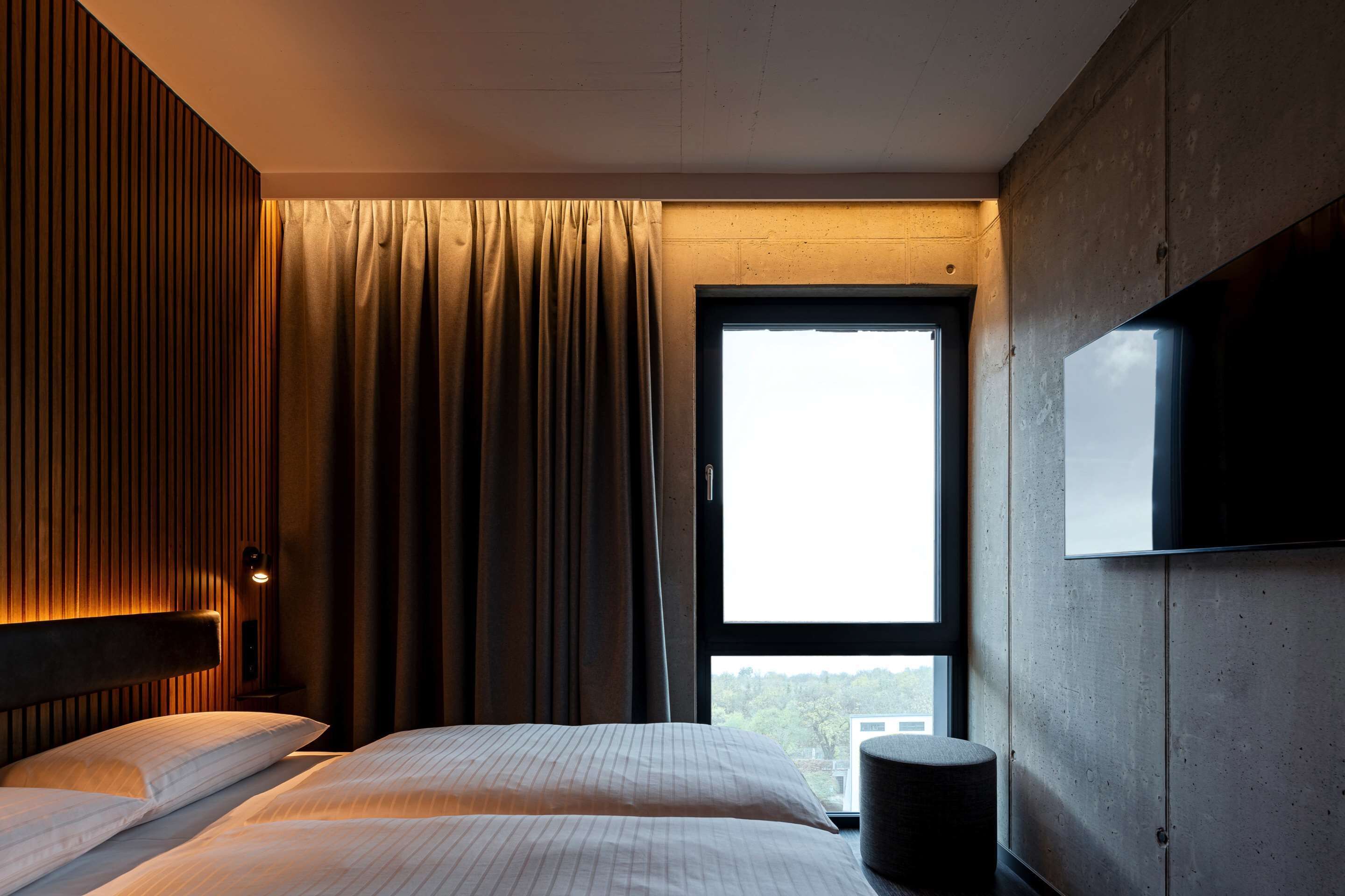 Una habitación de hotel con un ventanal grande, una cama con dos almohadas, una mesita de noche con lámpara y un televisor montado en la pared.