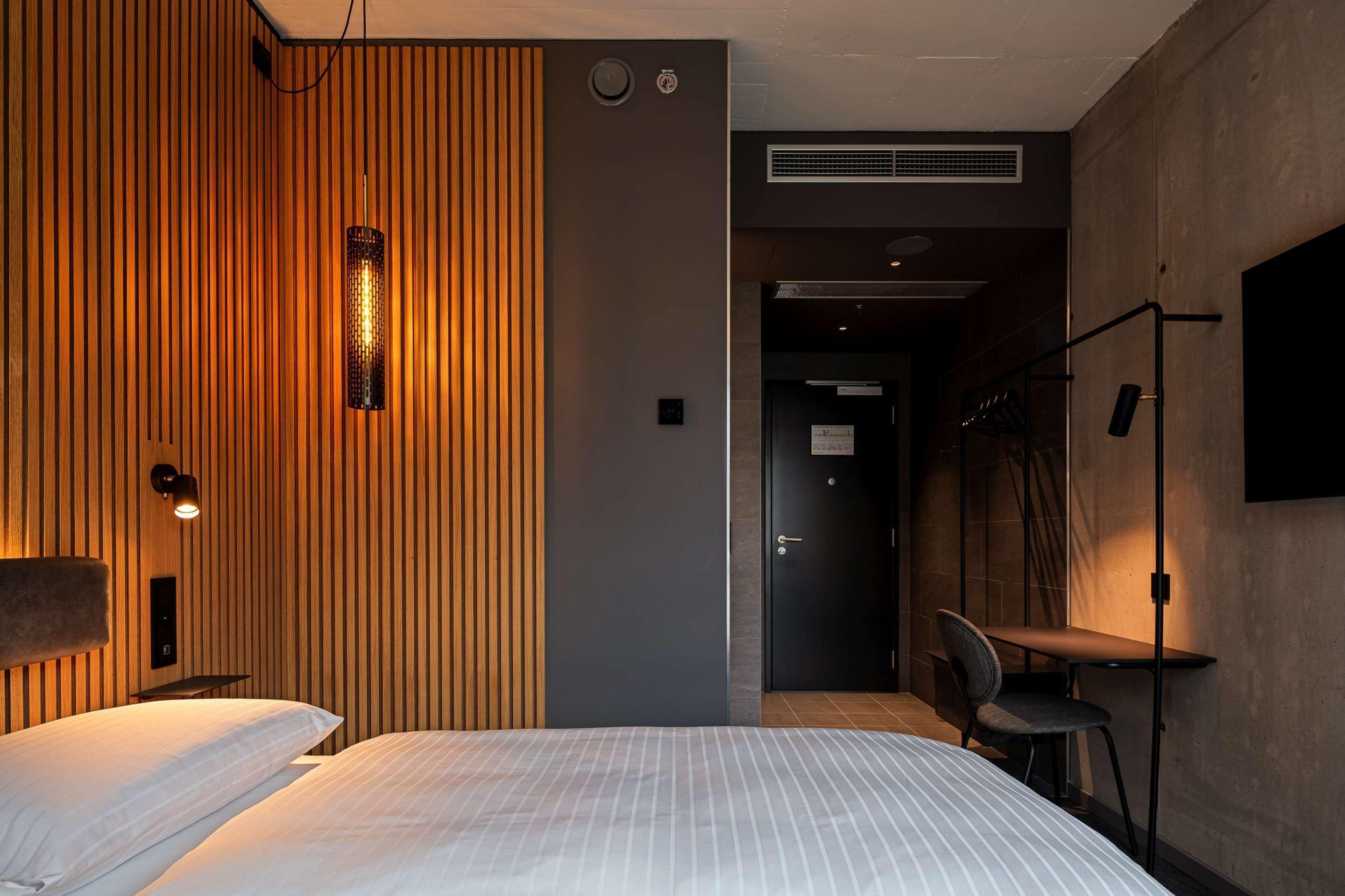 Una habitación de hotel moderna con una cama grande, un escritorio y una silla. Hay un televisor montado en la pared y una lámpara sobre la cama.