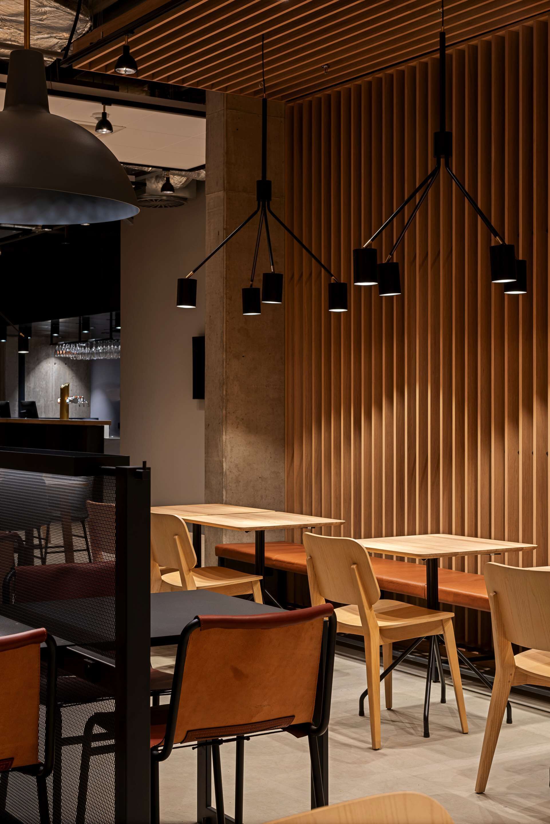 Un interior de restaurante moderno con mesas y sillas de madera, iluminación colgante y una zona de bar al fondo.